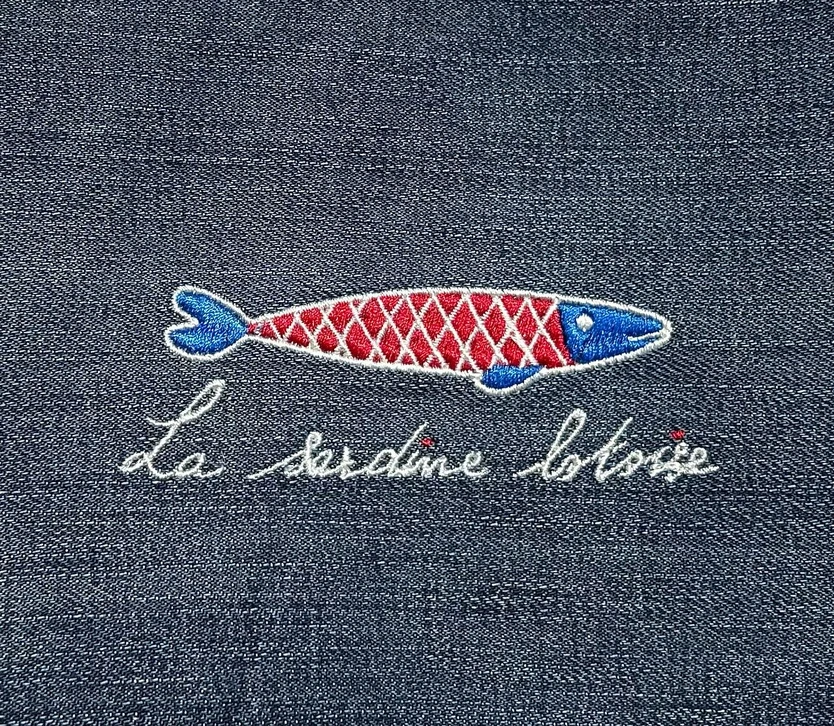 Photographie professionnelle en gros plan de plusieurs produits personnalisés de la marque La Sardine Lotoise disposés harmonieusement sur une surface neutre : un doudou brodé de couleur pastel, un accroche-tétine tissu personnalisé, un mug blanc avec un logo ou motif brodé discret, et un petit transat textile plié montrant sa personnalisation. Éclairage naturel et doux, mise en scène minimaliste pour mettre en avant la qualité et l'exclusivité des créations. Les produits doivent être reconnaissables et représentatifs de l'offre unique de la marque.