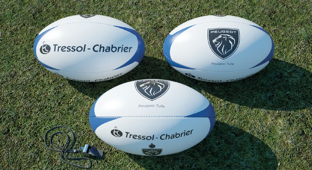 Gros plan détaillé montrant une composition de créations Archéon : un t-shirt ou sweat-shirt personnalisé avec broderie visible de qualité professionnelle (par exemple des motifs aux couleurs vives), un mug blanc avec décor imprimé en transfert DTF, et un accessoire textile promotionnel (comme un transat ou doudou Sardine Lotoise). Les produits sont arrangés de manière esthétique sur une surface neutre, mettant en avant la qualité des finitions, les détails de la broderie et l'impact visuel des personnalisations, photographiés en lumière professionnelle.