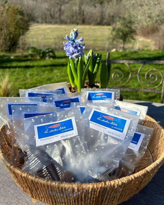 Photographie professionnelle en plan moyen montrant la table ou l'établi d'un atelier de personnalisation textile, avec des matériaux et fournitures visibles : bobines de fil de couleurs variées (rouge, bleu, or, blanc), carrés de tissu ou échantillons textiles, aiguilles, ciseaux de broderie, cercle à broder contenant un morceau de tissu en cours de travail, tous disposés de manière organisée et esthétique. Surface de travail en bois naturel clair, lumière naturelle ou professionnelle douce, fond légèrement flou montrant l'atelier en arrière-plan. L'image doit transmettre l'atmosphère artisanale, la rigueur et le professionnalisme du savoir-faire d'Archéon, mettant en avant les outils de la broderie et la préparation des créations sans montrer de personnes.