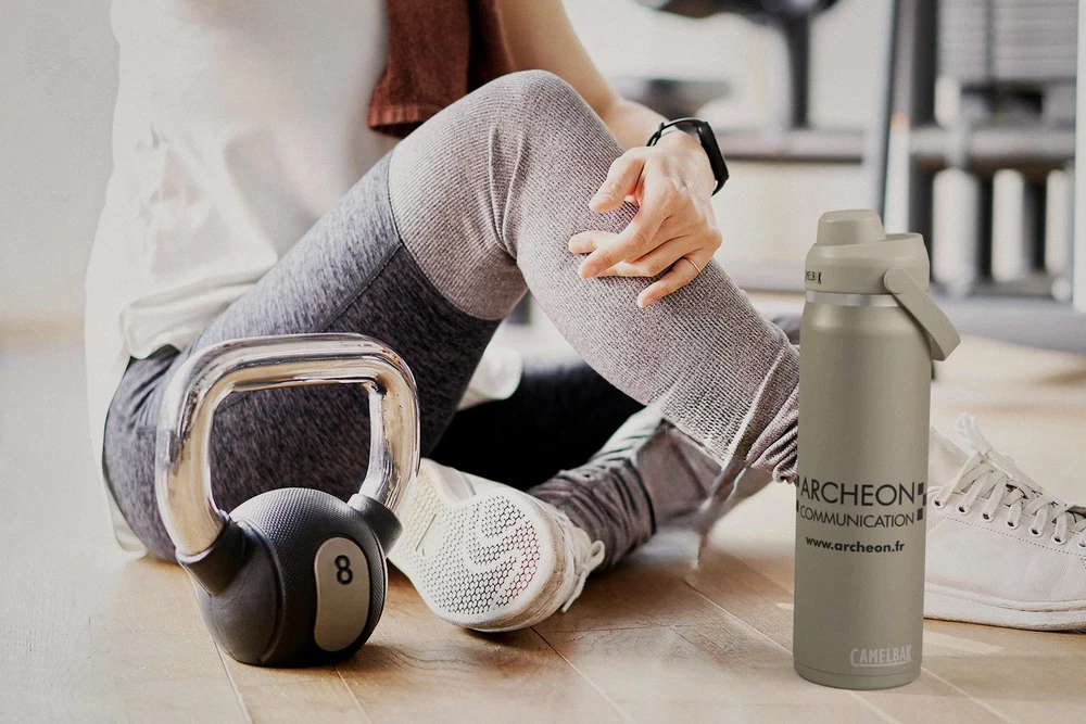 Photographie professionnelle en plan rapproché montrant une collection variée de produits personnalisés Archéon arrangés de manière esthétique : un polo ou chemisette en coton avec broderie détaillée (logo ou motif coloré en points nets), une casquette ou chapeau avec logo brodé en relief, une batterie externe ou accessoire informatique avec impression de qualité, le tout disposé sur une surface en bois naturel clair, sous lumière douce et professionnelle, mettant en évidence les textures, les finitions et la qualité des personnalisations. Fond neutre gris très clair. Cette image doit montrer clairement la diversité des produits promotionnels de l'entreprise et la qualité des techniques de personnalisation appliquées.