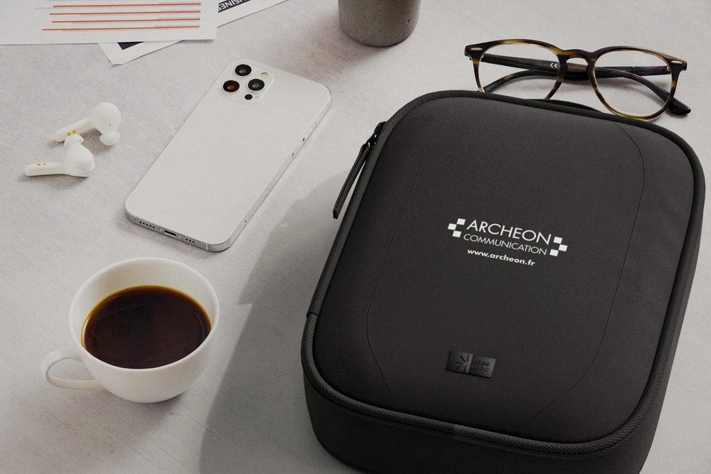 Photographie en plan large d'un atelier de personnalisation textile moderne, montrant un artisan en train de travailler sur une machine de broderie professionnelle avec un vêtement de marque (polo, t-shirt ou veste), sous éclairage naturel. L'arrière-plan affiche discrètement des échantillons de produits personnalisés exposés : textiles brodés, mugs, objets publicitaires colorés. L'atmosphère doit être professionnelle, sérieuse et refléter l'expertise artisanale et la qualité de contrôle d'Archéon.