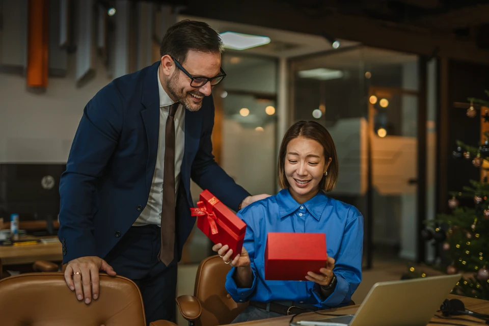 Jeune femme souriante tenant un cadeau emballé avec ruban rouge, posant dans une attitude réfléchie avec le doigt sur le menton, regard levé vers le haut