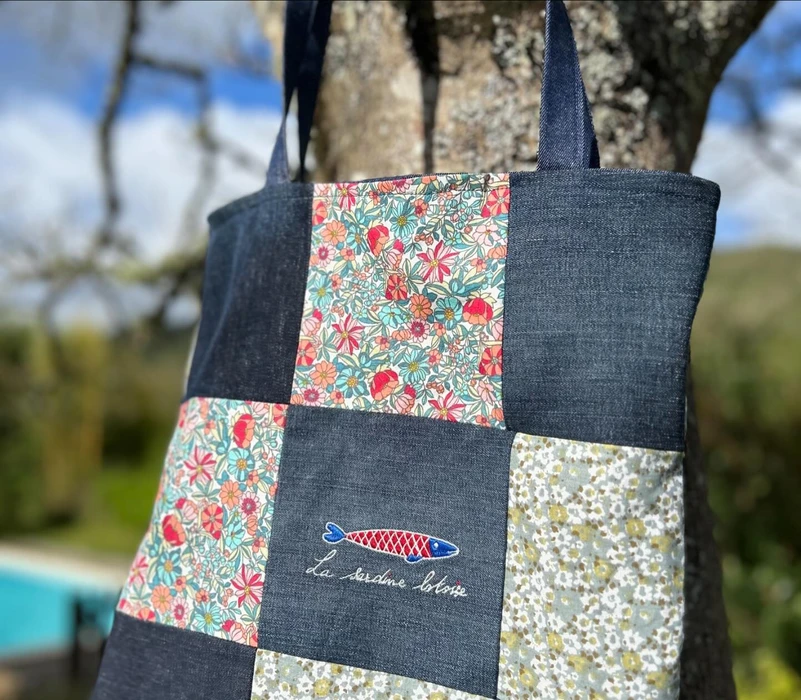 Photographie professionnelle en plan rapproché montrant un ensemble diversifié de créations Archéon disposées de manière dynamique et esthétique : un t-shirt ou sweat-shirt noir ou coloré présentant une broderie détaillée avec motifs éclatants, un mug blanc avec un design imprimé en transfert DTF visible et vibrant, et un accessoire promotionnel comme un transat plié ou un doudou de la marque Sardine Lotoise. Les produits sont arrangés sous différents angles pour mettre en valeur les textures, les finitions de qualité et les détails de personnalisation. Lumière naturelle ou professionnelle, fond neutre et minimaliste (gris clair ou blanc cassé) pour que les créations ressortent sans distraction.